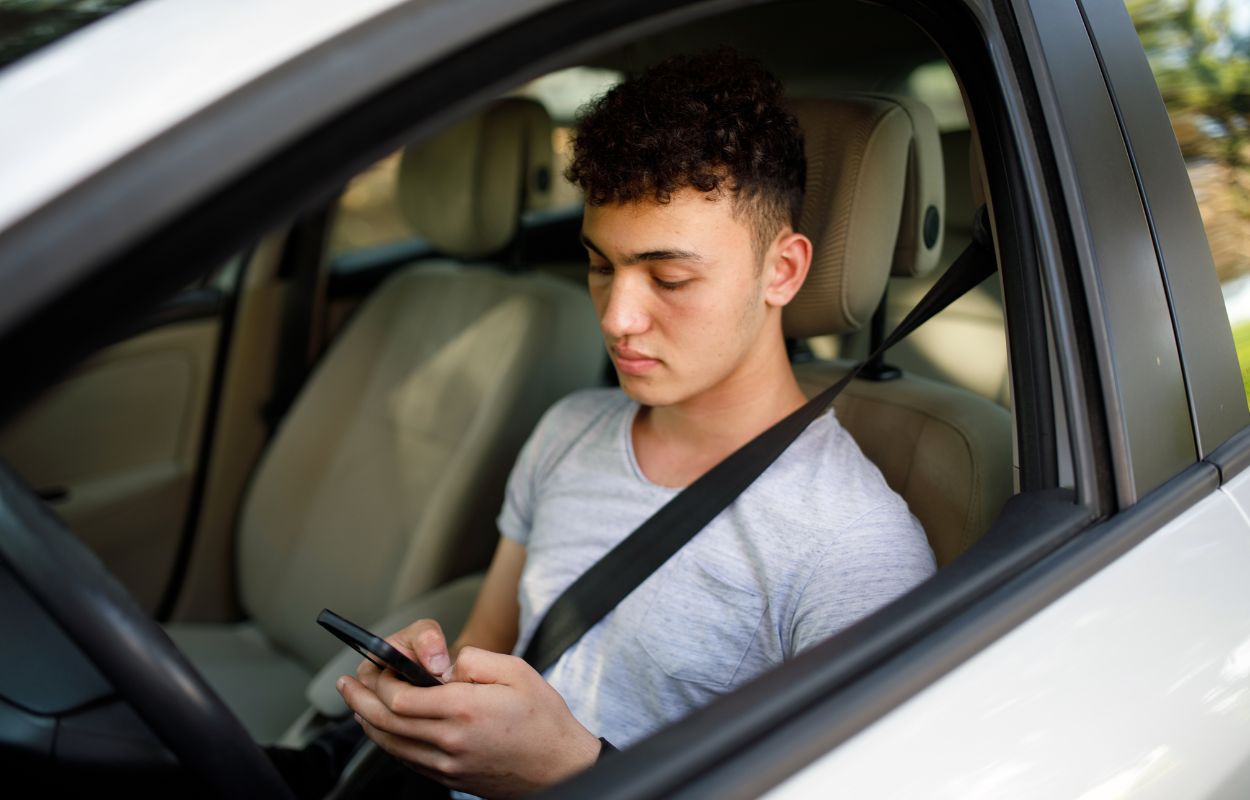 Young man sitting in driver's seat of white car, holding and staring at a cell phone.